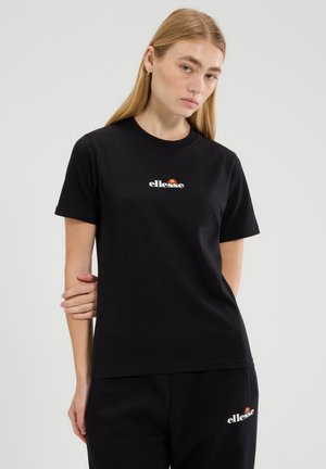 Mujer joven con cabello rubio liso, vistiendo camiseta negra de ellesse y pantalones a juego sobre un fondo claro y liso.