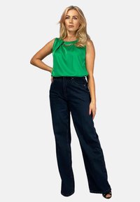 Zielony bezrękawnik z łańcuszkowym akcentem w połączeniu z ciemnoniebieskimi spodniami z szerokimi nogawkami; modelka stoi z jedną ręką na biodrze na białym tle.