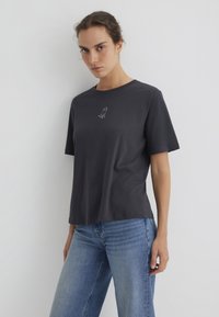 Dark gray cotton t-shirt with short sleeves, round neckline, featuring a small white dog embroidery on the chest. Paired with light blue jeans.