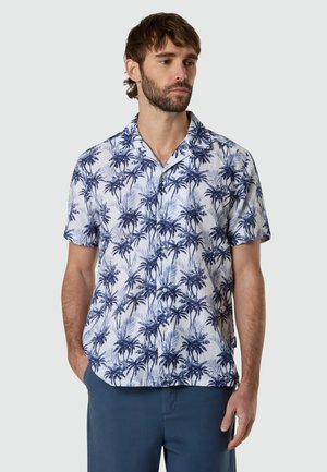 Mann trägt kurzärmeliges Hemd mit blauem Palmenmuster und dunkelblaue Shorts, steht mit einer Hand in der Tasche.