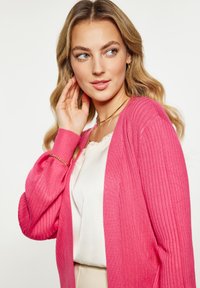 Ružový rebrovaný kardigán s dlhými rukávmi, nosený cez svetlú blúzku. Dizajn má voľný strih a textúrovanú látku, čo pridáva na jemnosti vzhľadu.