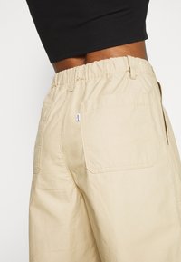Pantalon beige à taille élastique avec une coupe décontractée, poches latérales et une seule poche arrière avec des détails de coutures contrastées.
