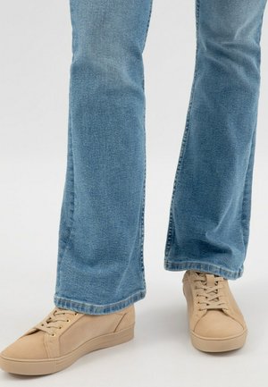 Beige Sneakers mit einem glatten Stoff-Obermaterial, runder Zehenpartie und flacher Gummisohle, kombiniert mit hellblauen, ausgestellten Jeans mit Denim-Oberfläche.