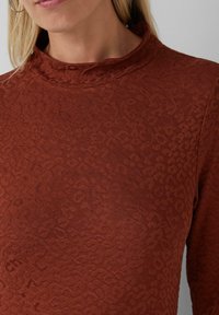 Femme portant un haut long à manches brunes texturé avec un col montant et un motif animal discret, mettant en valeur le cou et le haut du torse.