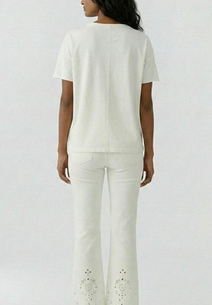 Vrouw met lang donker haar, gekleed in een effen wit T-shirt met korte mouwen en een witte broek met patroon uitsnijdingen bij de zomen, gezien van achteren.