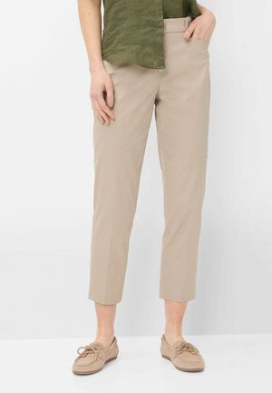 Person iført beige kortbukser, brune mokkasin-sko og en grøn skjorte, stående med den ene hånd i lommen mod en hvid baggrund.