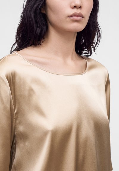 Femme aux cheveux foncés portant un chemisier en satin beige brillant avec un large col arrondi sur un fond clair uni.