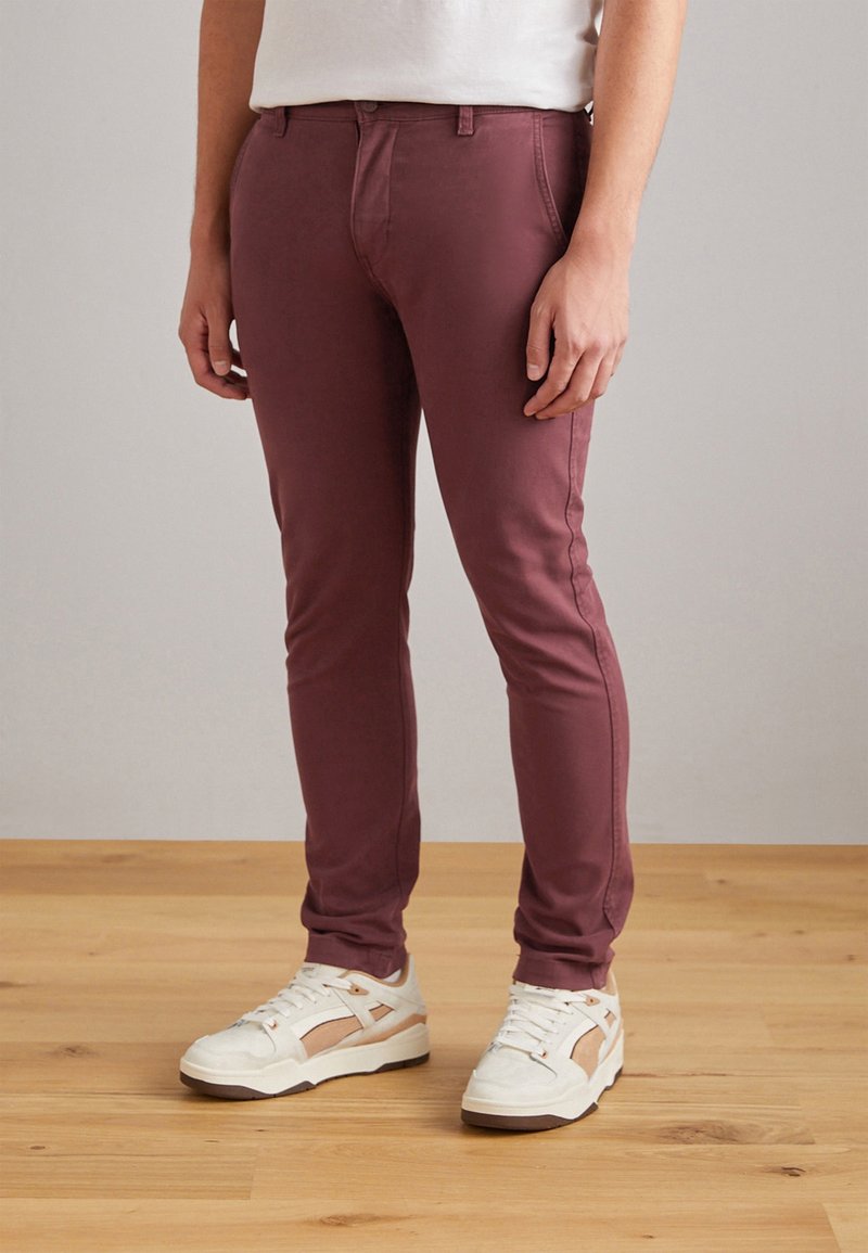 Homme portant un pantalon slim bordeaux et des baskets blanches, debout sur un sol en bois devant un fond uni.