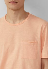 Licht perzikkleurig katoenen t-shirt met een ronde hals, korte mouwen en een klein borstzakje. Zachte textuur met een subtiele, uniforme afwerking.