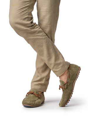 Person trägt beigefarbene Hose und olivgrüne Mokassins aus Wildleder mit braunen Schnürsenkeln, steht mit gekreuzten Beinen vor weißem Hintergrund.