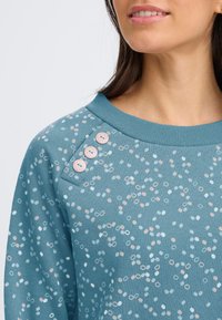 Felpa azzurro chiaro con un motivo strutturato di cerchi di dimensioni diverse in bianco e rosa, con tre bottoni decorativi sulla spalla.