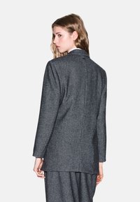 Blazer grigio a spina di pesce con taglio sartoriale, maniche lunghe e dettaglio di apertura sul retro. Realizzato in tessuto testurizzato, mostra un design classico.