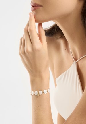 Woman wearing white sleeveless top touching chin with left hand adorned by white circular bead bracelet with small gold charm.