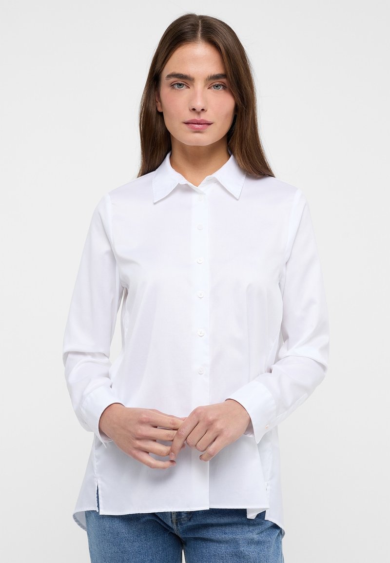 White button-up shirt with a classic collar, long sleeves, side slits, and a smooth texture. Paired with blue jeans.