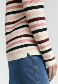 Juostuota megztinis, pagamintas įvairių rožinės, burgundiškos ir juodos spalvų atspalvių, su tekstūruota medžiaga ir šoninėmis įplautomis, derinamas su džinsais juosmens srityje.