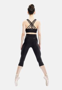 Woman in black crop top and leggings performing ballet en pointe with legs apart, hair in bun, facing away on plain white background.