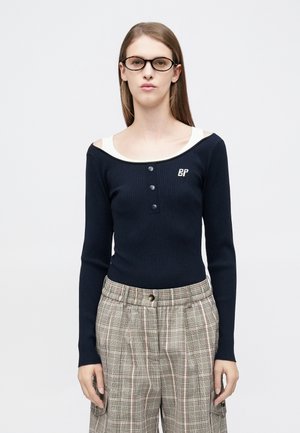 Maglia a maniche lunghe a coste blu marina con ampia scollatura color avorio, patta con tre bottoni e piccolo logo "BP" sul petto. Abbinata a pantaloni a quadri.