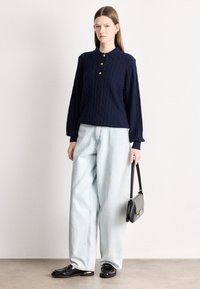 Μπλε ναυτικό πουλόβερ με πλεκτό σχέδιο και τρία χρυσά κουμπιά, ανοιχτό μπλε τζιν με φαρδύ πόδι, μαύρα μοκασίνια και μια μικρή μαύρη τσάντα με χρυσή λεπτομέρεια.