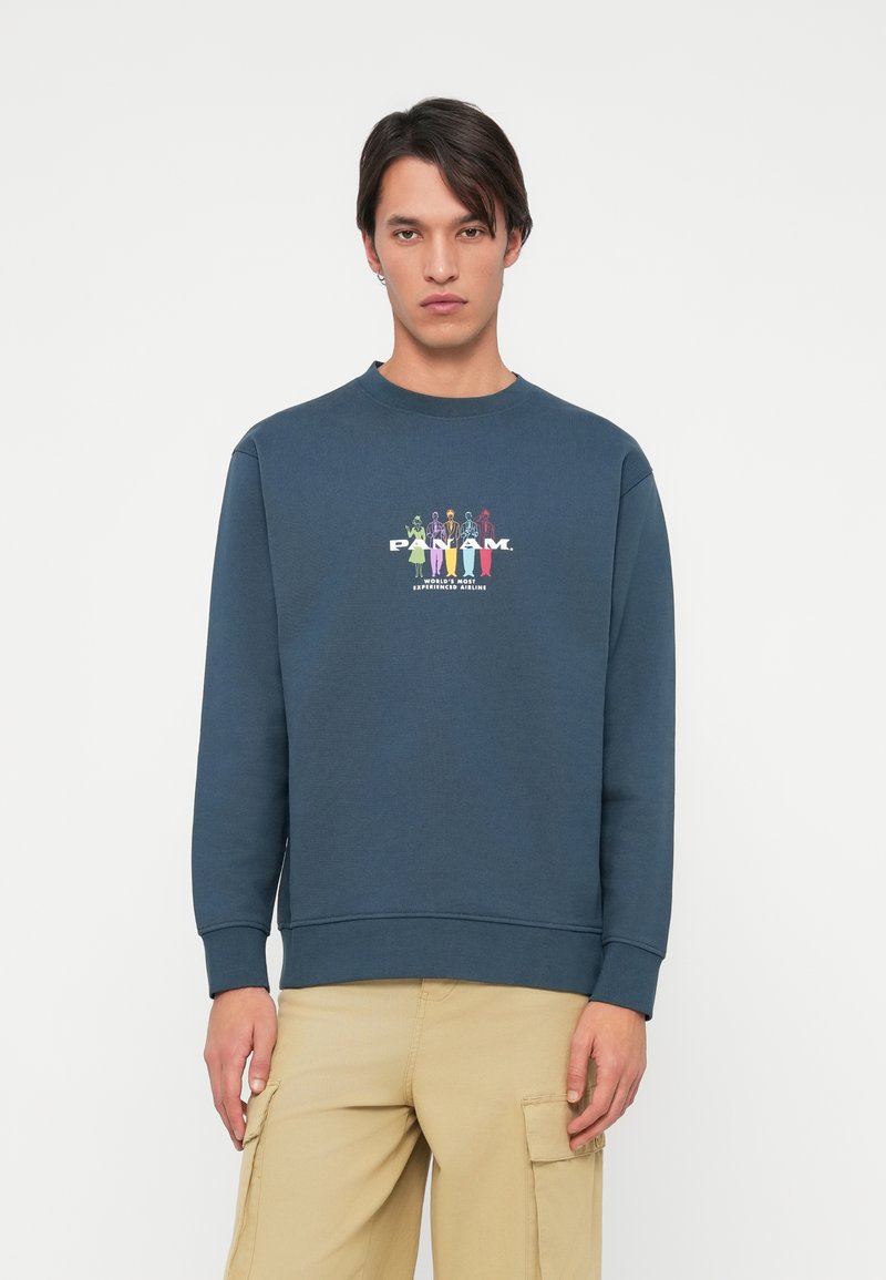 Marineblauwe sweatshirt met een ronde hals, geribde manchetten en zoom. Voorzien van een kleurrijke grafische afdruk van figuren en de tekst "PAN AM" op de voorkant.