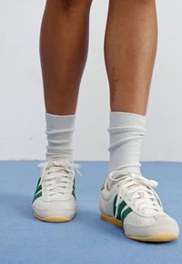 Tênis esportivos brancos com riscas verdes, detalhes perfurados e solas bege, combinados com meias de cano curto brancas e caneladas.