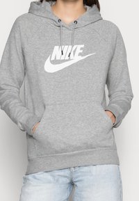 Person som bär en ljusgrå Nike-hoodie med vitt logo, händer i framfickan, kombinerad med ljusblå jeans, endast överkropp synlig.
