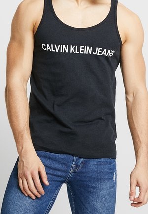 Mężczyzna w czarnej koszulce bez rękawów z napisem „CALVIN KLEIN JEANS” i niebieskich dżinsach, stoi ze swobodnie opuszczonymi ramionami.