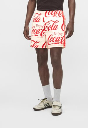 Mężczyzna w beżowych szortach z czerwonym wzorem logo Coca-Cola, białych skarpetkach i kremowych trampkach z czarnymi paskami.