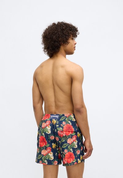 Jeune homme aux cheveux bouclés portant un short de bain bleu marine orné d'un imprimé floral rouge et rose, vu de dos sur un fond uni.