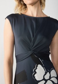 Dunkelblaues Kleid mit kurzen Ärmeln, das ein geknotetes Taillendetail und ein großes weißes Blumenmuster aufweist. Glattes, leichtes Material.