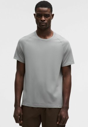 Mężczyzna w prostym jasnoszarym koszulce z krótkim rękawem i ciemnych spodniach, stojący na jednolitym białym tle.