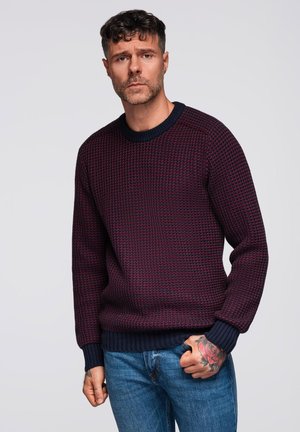 Pull en laine de couleur bordeaux avec un motif texturé et des poignets et un ourlet côtelés en bleu nuit. Porté avec un jean bleu, offrant une coupe décontractée.