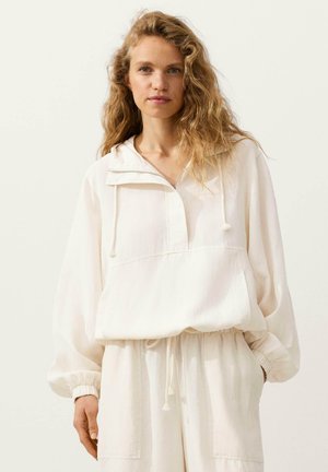 Vrouw met golvend blond haar die een losse, crèmekleurige trui met halve rits en bijpassende broek met trekkoorden draagt, staand tegen een effen achtergrond.