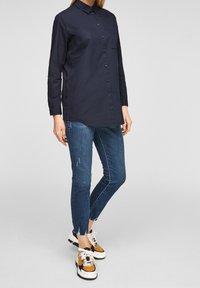 Mörkblå skjorta med långa ärmar och bröstficka, i kombination med slitna blå smala jeans och flerfärgade sneakers.