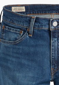 Jeans Levi's en denim bleu avec poches avant, passants pour ceinture, rivets métalliques et une étiquette Levi's Premium visible à l'intérieur de la ceinture.