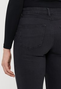 Juodai džinsai su prigludusia siluetu, turintys dvi galines kišenes ir siuvinėjimo detales. Audinys turi lygų paviršių ir šiek tiek blizgesio.