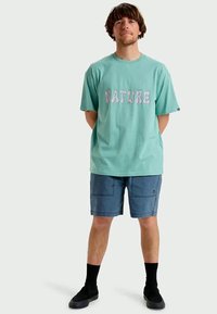 T-shirt de algodão turquesa com o texto estilizado "NATURE" em violeta claro. Combinado com calções azuis e meias pretas. Corte casual e mangas curtas.