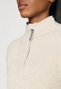 Pull-over crème à col montant, en tissu tricoté texturé, doté d'une fermeture à zip demi-longueur et d'une finition de cou lisse. Sensation douce et chaleureuse avec des coutures détaillées.