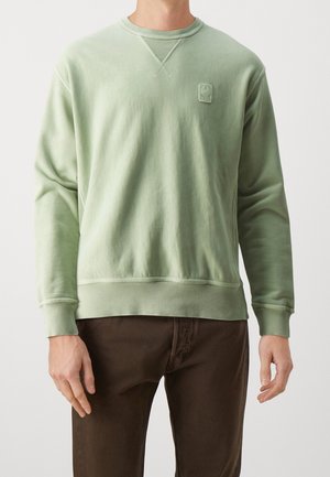 Mann trägt hellgrünes langärmliges Sweatshirt mit kleinem Brustlogo und braune Hose vor einfachem hellem Hintergrund.