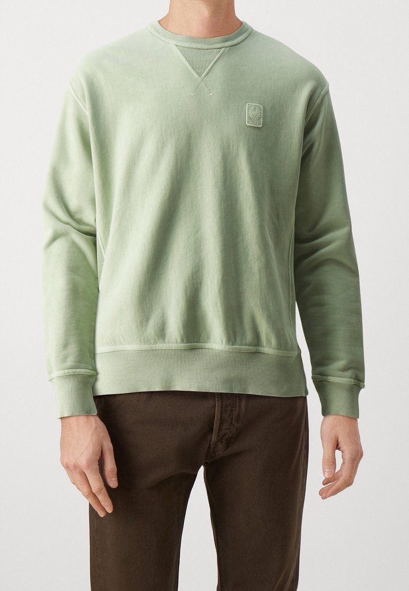Homme portant un sweat-shirt à manches longues vert clair avec un petit logo sur la poitrine et un pantalon marron sur un fond clair uni.