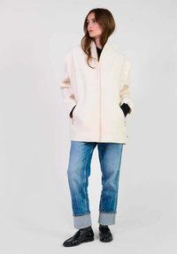 Crème-kleurige wollen jas met hoge kraag, verlaagde schouders en voorkruiden; gecombineerd met omgeslagen blauwe denimjeans en zwarte schoenen.