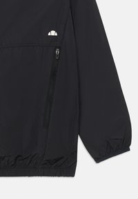 Svart vindjacka med en framficka med dragkedja, elastiska ärmslut och nederkant. Har en liten vit logotyp på övre bröstet. Lätt material.