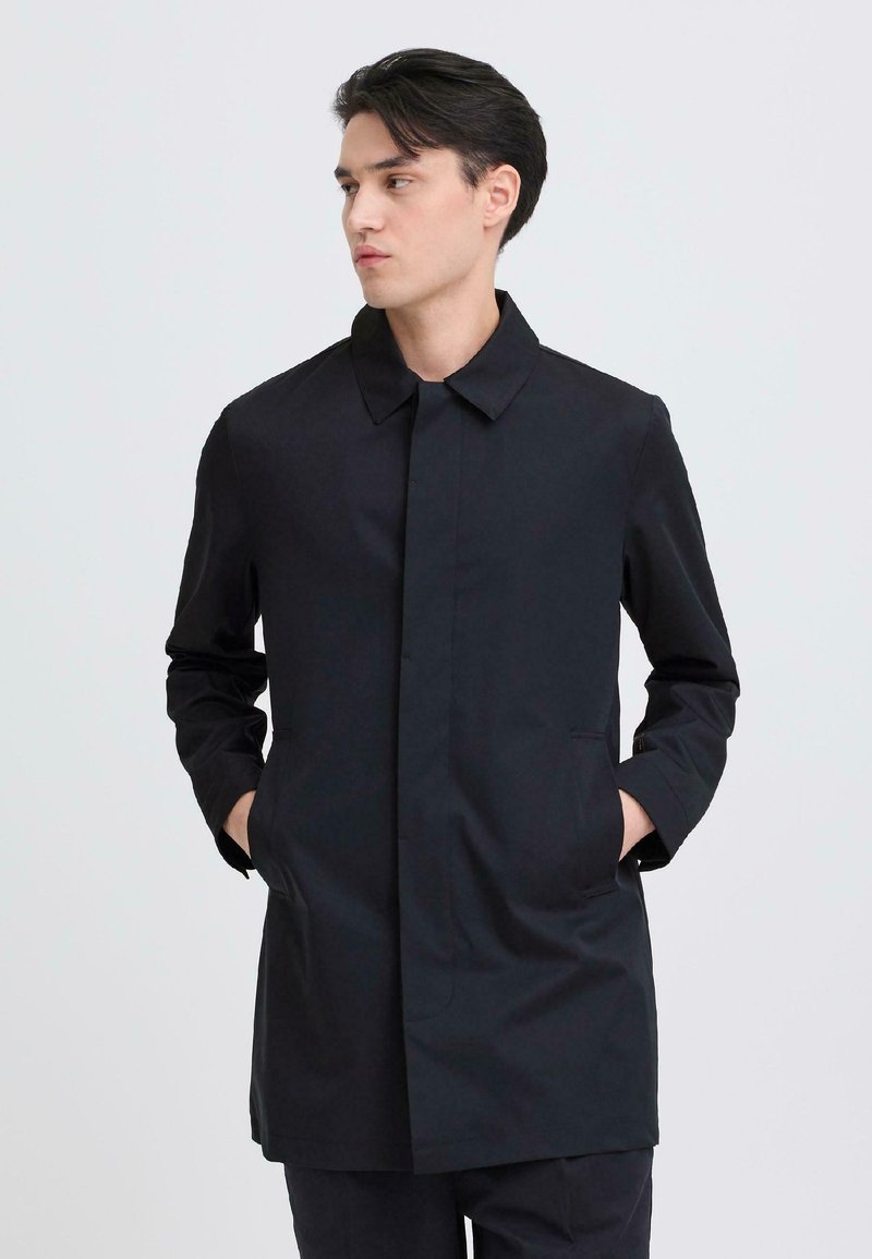 Mand iført en sort skræddersyet frakke med hænderne i lommerne, kigger mod venstre foran en ensfarvet lys baggrund.