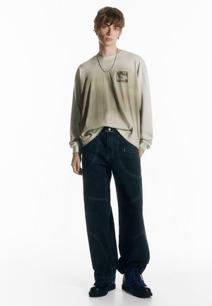 Cămașă cu mâneci lungi, bej deschis, cu patch grafic și model detaliat pe mânecă. Asortată cu blugi largi, bleu închis și cizme negre.