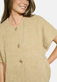Maglione a maniche corte beige in maglia con motivo a spina di pesce, dotato di grandi bottoni rotondi e una vestibilità morbida. Texture soffice e design leggero.