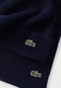 Tamsiai mėlynas megztas šalikas ir kepurė, abu su žaliu krokodilo logotipu ir raudonomis burnomis, išsiūtais ant audinio.
