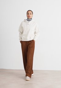 Hanorac gri deschis cu buzunar frontal și șnur, purtat peste un pulover cu guler înalt în dungi alb-negru. Pantaloni maro cu croială largă și adidași.