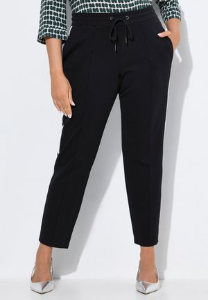 Donna che indossa pantaloni neri a sigaretta con cintura elasticizzata, una mano in tasca, abbinati a tacchi a punta argentati e a una maglia a maniche lunghe con motivo.