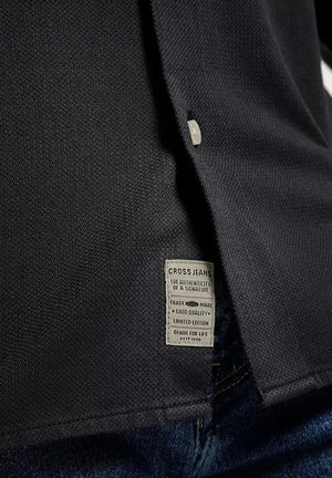 Czarna tkanina teksturowa z guzikiem i wszytą metką z napisem "CROSS JEANS", wskazującą markę i informacje o edycji limitowanej.