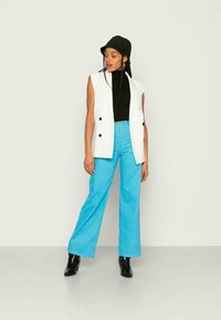 Blazer branco sem mangas com botões pretos, combinado com um top preto e calças largas azuis. Um chapéu preto e botas de tornozelo completam o visual.