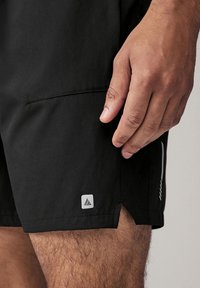 Shorts de sport noirs avec une texture lisse, comportant un petit logo blanc sur la cuisse gauche et des poches latérales pour le rangement.